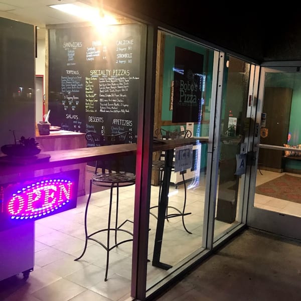 Exterior view of Bobo's Pizza at night, with a glowing "OPEN" sign and menu boards visible through the window.